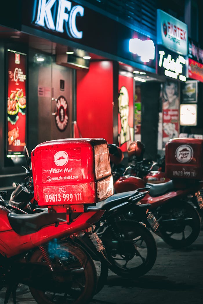 Pizza delivery motorbikes parked outside KFC and Pizza Hut in urban Dhaka at night.